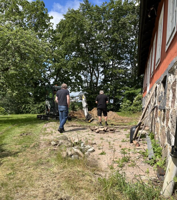 Två personer står vid ett hus och arbetar med att gräva upp marken för att sätta ner plintar, maskiner och stenar syns i bakgrunden.