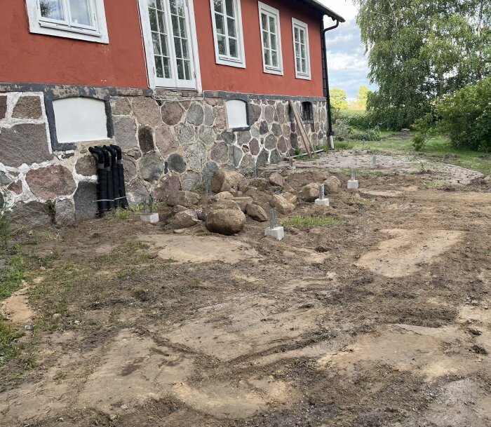 Bild av framtida balkongområde med stenhusvägg i bakgrunden. Plintar och antal stora stenar synliga på marken efter grävarbete är avslutat.