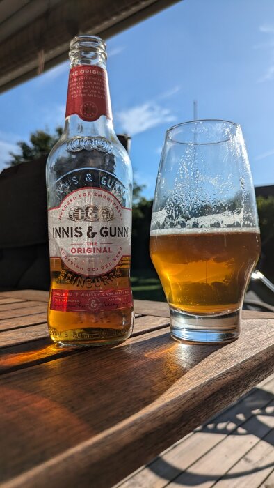 Flaska och glas med öl från Innis & Gunn står på ett träbord utomhus under en solig dag.