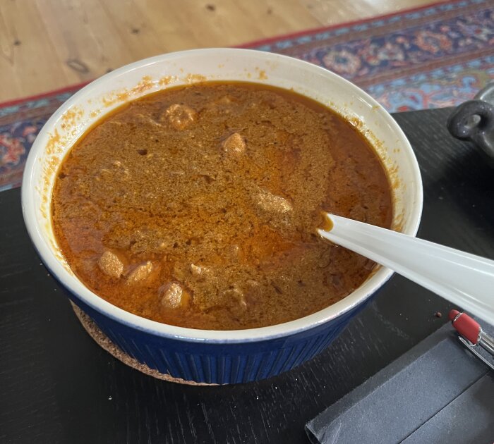 En skål med färdiglagad butter chicken ståendes på ett svart bord, med en vit sked i skålen.