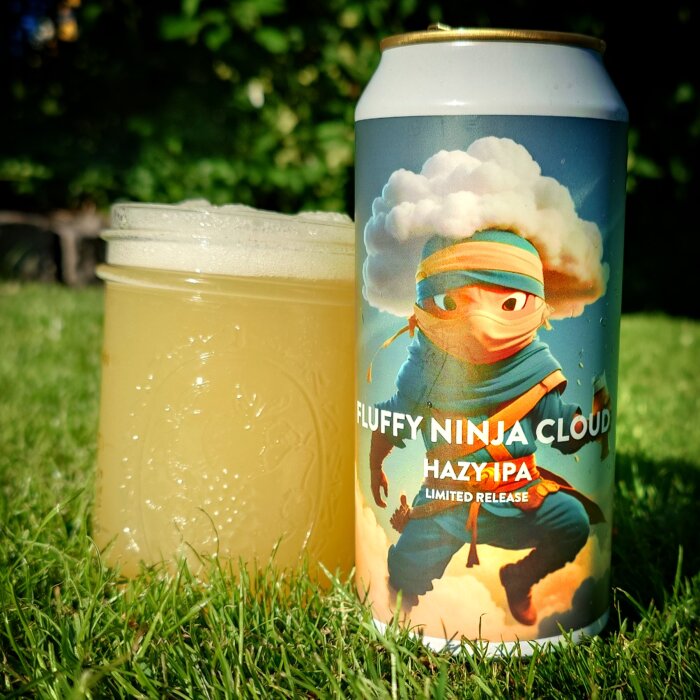 En burk med ölen "Fluffy Ninja Cloud Hazy IPA" står på gräset bredvid ett glas med öl.