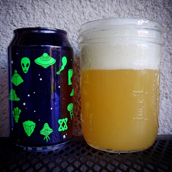 En burk Omnipollo Zodiac IPA med rymdmotiv står bredvid ett glas med skummig öl på ett galler.