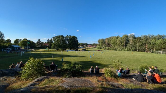 Fotbollsmatch mellan Stegeborgs IF och Nyköpings BIS på en gräsplan, omgiven av träd och sittande åskådare i förgrunden. Klarblå himmel.