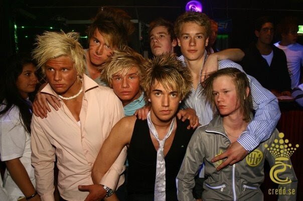 En grupp unga människor poserar för ett foto på en nattklubb, med färgstarka frisyrer och utstyrslar, potentiellt relaterade till en diskussion om en kommun i Täby.