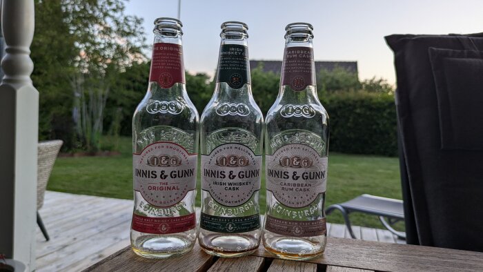 Tre tomma ölflaskor av märket Innis & Gunn står uppradade utomhus på ett träbord, omgiven av gräsmatta och trädgård i bakgrunden.