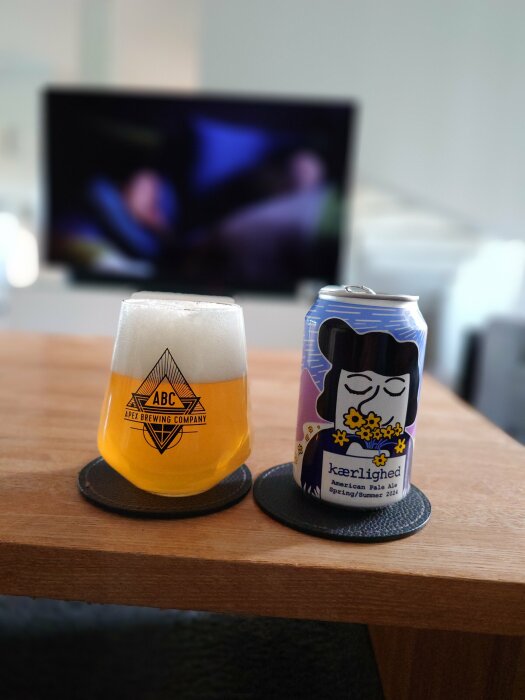 Ett ölglas från Apex Brewing Company och en burk med "kærlighed" American Pale Ale står på ett bord framför en suddig TV-bild.