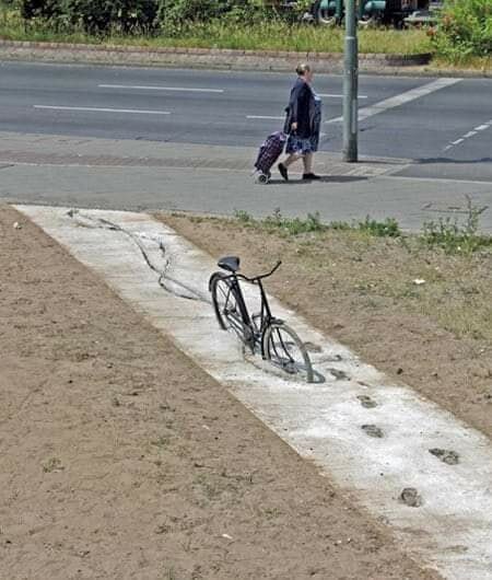 Omkullfallen cykel liggandes på en gångväg, med tydliga spår efter hjulet i den våta betongen. En person går förbi på trottoaren längre bort.