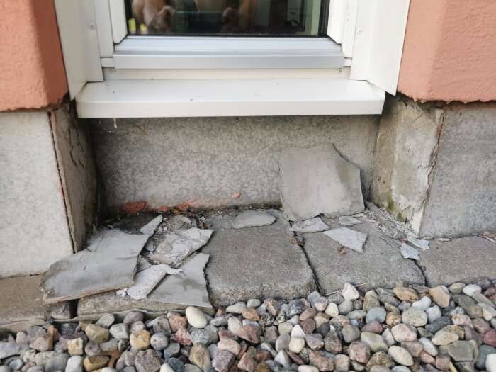 Skadat och söndrat cement vid botten av ett fönster, med stenläggning och småsten framför.