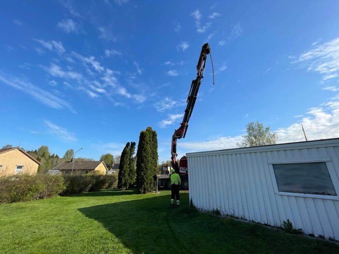 En person övervakar en kran som lyfter ett spabad på plats bredvid en vit byggnad i en trädgård, med blå himmel och några träd i bakgrunden.