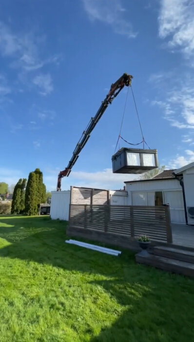 En kran lyfter ett spabad på plats vid en altan under en klarblå himmel. Grönt gräs och träd syns i bakgrunden.