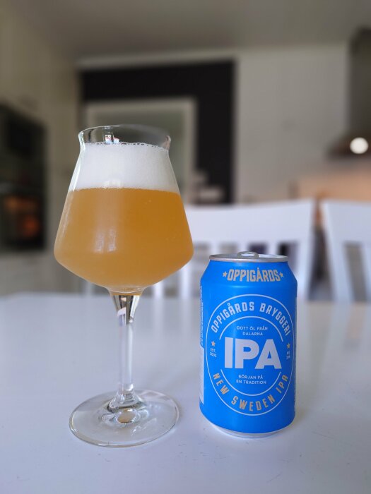 Ett glas med guldfärgad Oppigårds New Sweden IPA-öl bredvid en blå burk med samma märkesetikett på ett vitt bord i en köksmiljö.