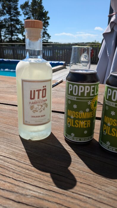 En flaska Utö ekologisk snaps och två burkar Poppels Midsummer Pilsner står på ett träbord utomhus, med en pool och skog i bakgrunden.