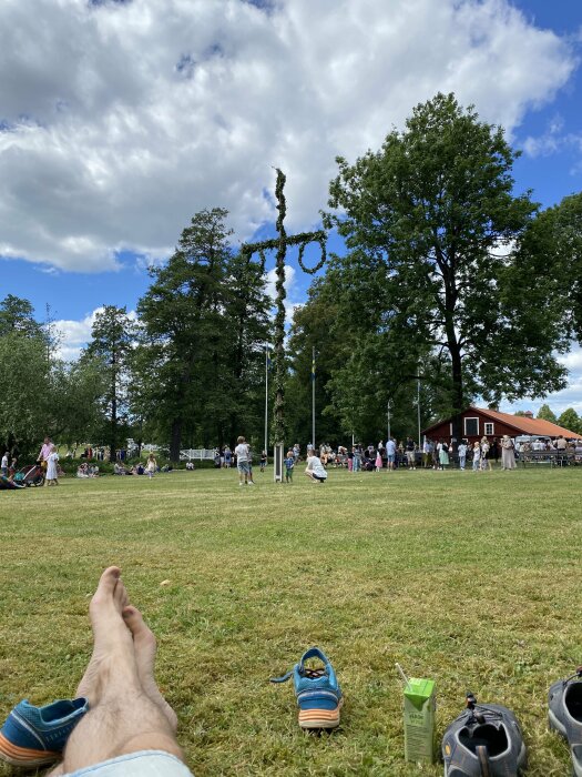 En grön midsommarstång på en gräsplan, människor firar med folkmusik och lekar, blå himmel med vita moln, ett par fötter och skor i förgrunden.