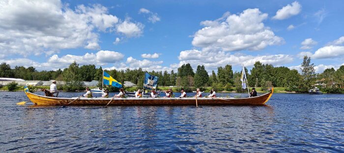 Personer i traditionella kläder paddlar en lång träbåt på en sjö. Svenska flaggor och andra flaggor syns. Bakgrunden visar skog och några byggnader.