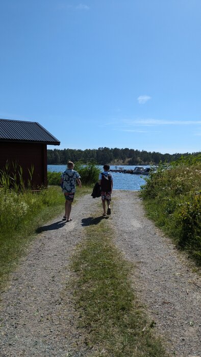 Två personer går på en grusväg mot en sjö med en brygga och båtar på en solig dag, med gräs och en röd byggnad på sidorna.