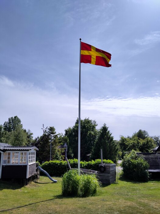 En skånsk flagga vajande på en flaggstång i en trädgård med ett lekhus, rutschkana och grönska i bakgrunden under en klarblå himmel.