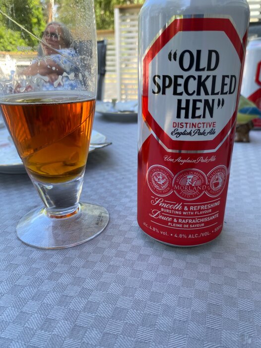 Burk med "Old Speckled Hen" engelsk pale ale och ett halvtomt ölglas står på ett bord, suddig bakgrund med någon som sitter ute i solen.