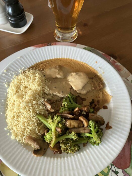 En maträtt med fläskytterfilé i sås, stekt broccoli och svamp, samt couscous på en vit tallrik. I bakgrunden syns ett glas öl.