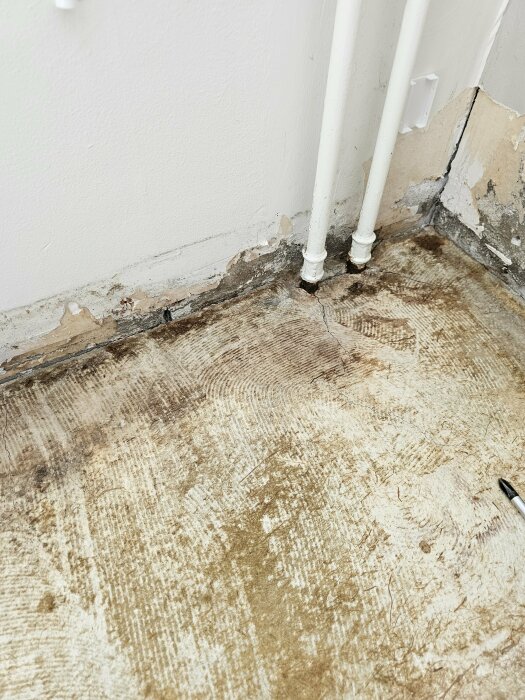 Hörn av ett rum med avskavd väggputs nära två vita rör. Synlig spricka längs golvet.