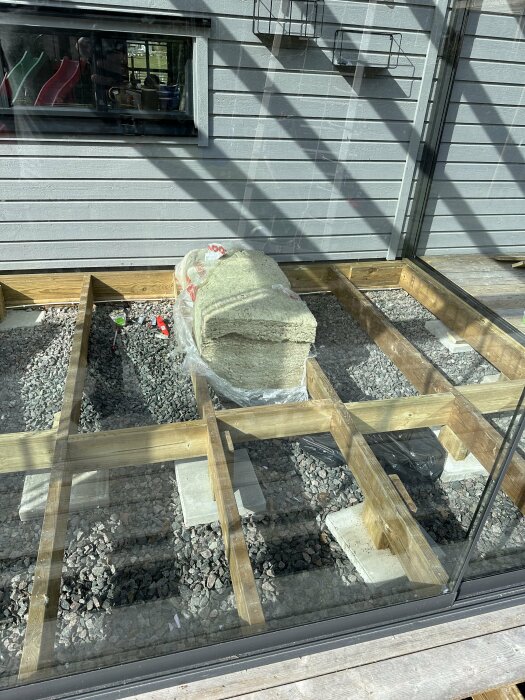 Rockwoolisolering på ett grusunderlag inom ett ramverk av träreglar, färdigställt för installation under ett trädäck, sedd genom ett glasfönster.