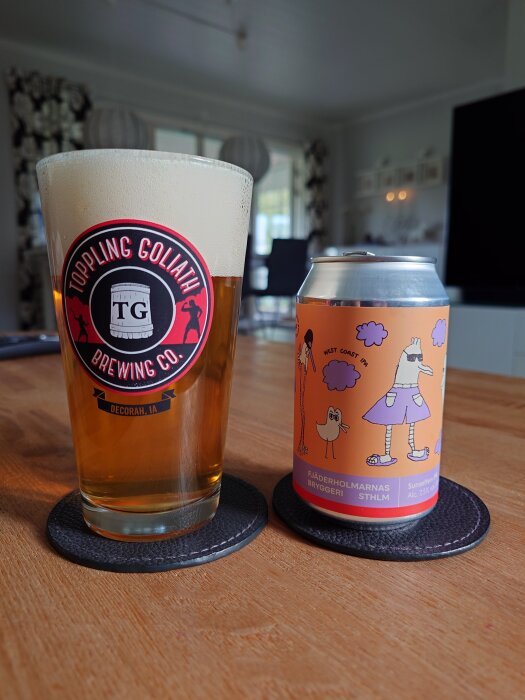 Ett glas öl med texten "Toppling Goliath Brewing Co." bredvid en ölburk från Fjäderholmarna Bryggeri med tecknad etikett, ståendes på ett träbord.