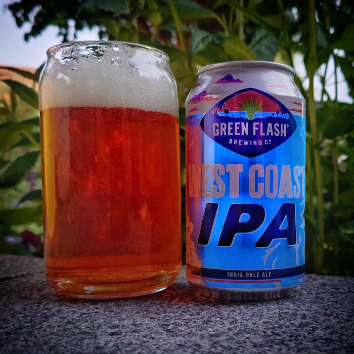 Öl i glas bredvid burk av märket Green Flash West Coast IPA, placerat utomhus med grönska i bakgrunden.