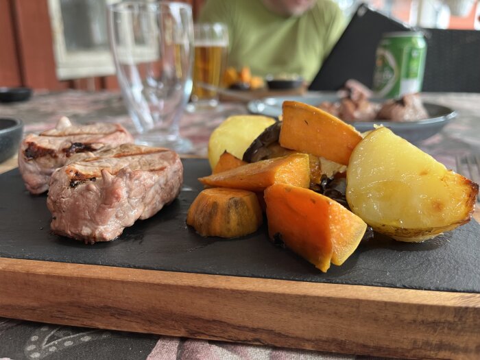 Grillad fläskfilé och ugnsstekta grönsaker, inklusive sötpotatis och färskpotatis, serverad på en tallrik med en ölburk och glas i bakgrunden.