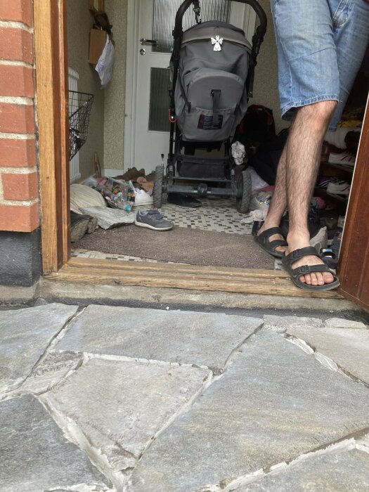 Bild av en dörrtröskel där tröskeln är högre än stenplattorna utomhus. En man i shorts och sandaler står i dörröppningen, och en barnvagn syns inomhus.
