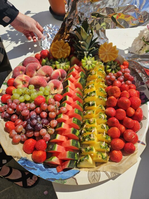 Fruktfat med skivor av kiwi, ananas, vattenmelon, persikor, druvor och jordgubbar. En hand håller folie i bakgrunden.