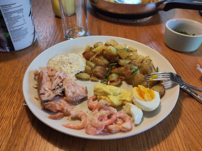 Tallrik med rökta laxrester, räkor, egengjord romsås, aioli, stekt färskpotatis och en halva kokt ägg på ett träbord.