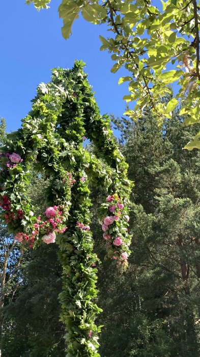 En midsommarstång dekorerad med gröna blad och blommor står utomhus på en solig dag med blå himmel.