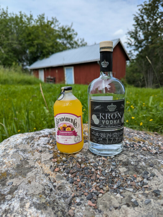 En flaska Bundaberg passionfrukt läsk och en flaska Kron vodka står på en sten med en röd stuga i bakgrunden.