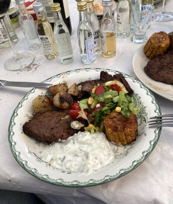Entrecôte med potatis, sallad, majs och tzatziki på en tallrik, omgiven av olika dryckesflaskor och ett glas rödvin på ett matkreativt midsommarfirande.