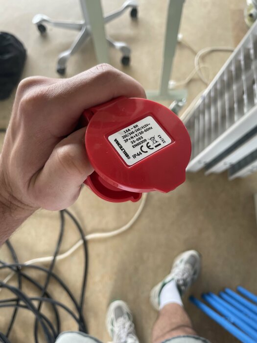 Närbild på en hand som håller en röd 16A kontakt med en etikett som visar tekniska specifikationer. I bakgrunden syns en svart kabel och delar av ett rum med möbler och utrustning.