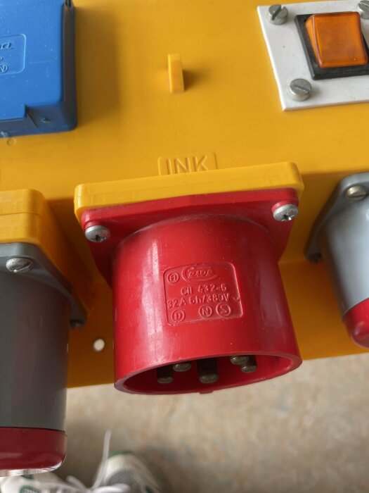 Byggcentral med en röd 32A, 6h, 380V kontakt (hane) från märket PCE, omgiven av flerfärgade uttag, på en gul bas märkt “INK”.