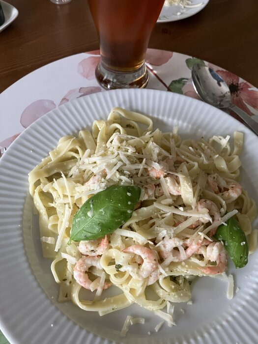 Pasta med räkor, basilika, parmesan och sås av fänkål, vitlök och rödlök på en vit tallrik, med en kopp mörk dryck i bakgrunden.
