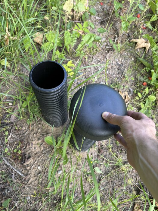 En person håller ett svart plastlock bredvid två svarta ihåliga rör stående vertikalt i gräs.