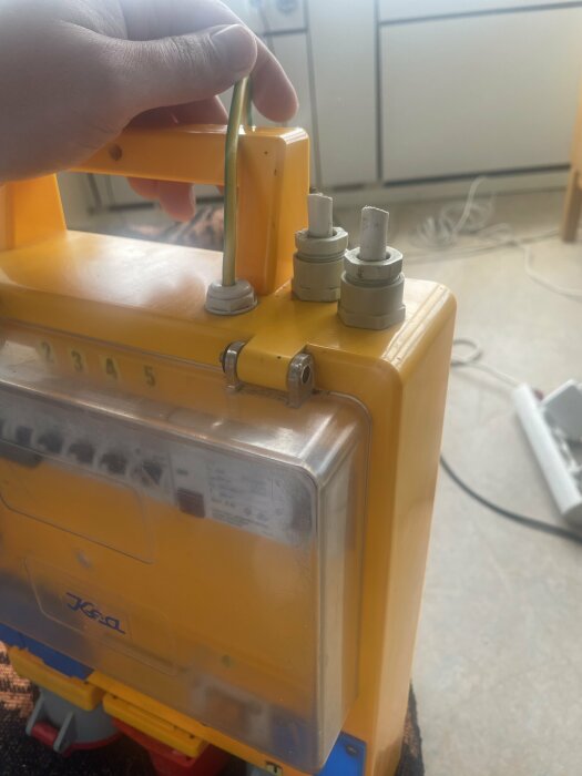 Gul elkapsling med en avkapad vit kabel och en hand som håller en gröngul jordkabel. Bakgrunden visar en inomhusmiljö med golv och möbler.