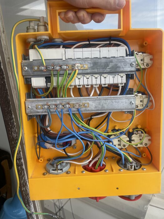 Gul elcentral med öppet lock som visar flera färgglada kablar anslutna till olika brytare och kopplingar. Hand håller i handtaget på toppen.
