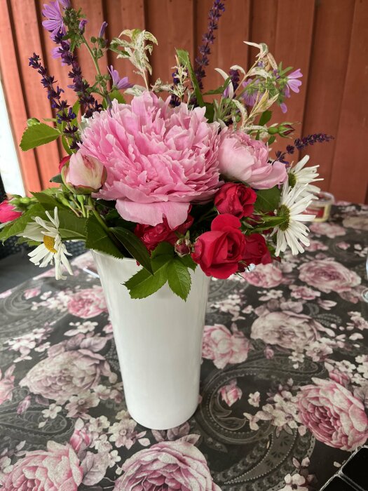 En vit vas med en bukett blommor, inklusive rosa pioner, röda rosor, vita prästkragar och lila blommor, står på ett bord med en blommig duk framför en trävägg.