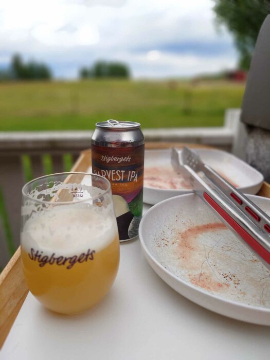 Öl från Stigbergets bryggeri i glas och burk bredvid tallrikar och grilltång, med suddig vy över ett fält i bakgrunden.