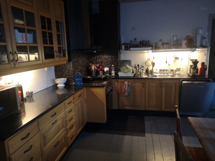 Kök i ett 1920-tals hus med träskåp, mörka bänkskivor, trägolv, diskbänk, och köksredskap. Mörk mosaikvägg bakom spisen och belysning under skåpen.