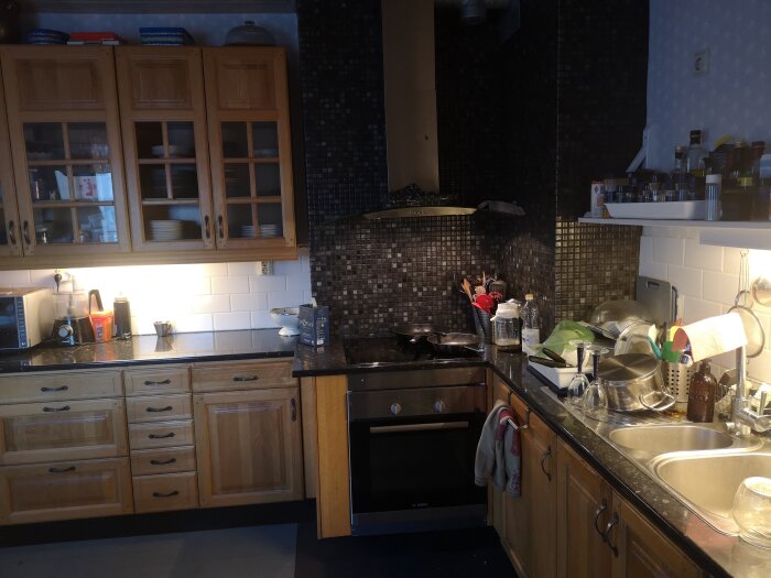 Kök med träskåp, svart kakel bakom spis och diskho, rostfri ugn och olika köksredskap. Hörn med murstock syns på bildens högra sida.