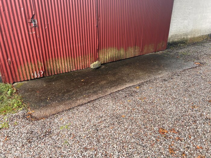 Röd plåtport med sliten yta och mossväxt. Vid portens fot syns en betongplatta, grus samt en sten. Marken är fuktig efter regn.