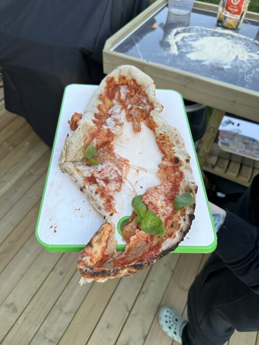 Misslyckad pizza med bränd och deformerad deg visas på en skärbräda utomhus, med ett bord med mjöl och en pizzaugn i bakgrunden.
