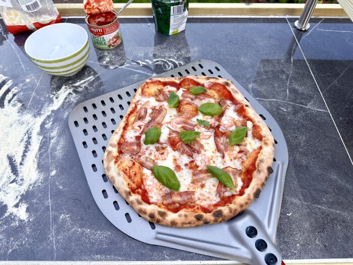 En nylagad pizza med skinka och basilika på en bakspade, bredvid en skål, pizzassåsburk och mjöl på en marmorbänk utomhus.
