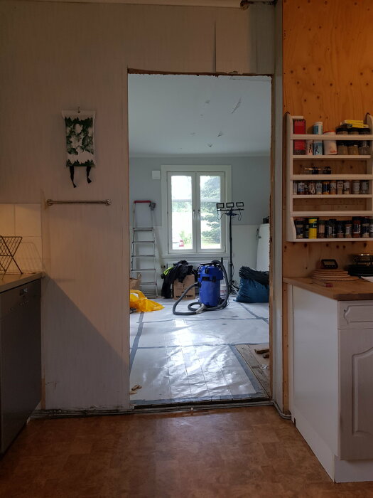 Renovering av kök med dörröppning utan dörr mellan kök och finrum, verktyg och material på golvet, dammsugare och stege synliga i finrummet.