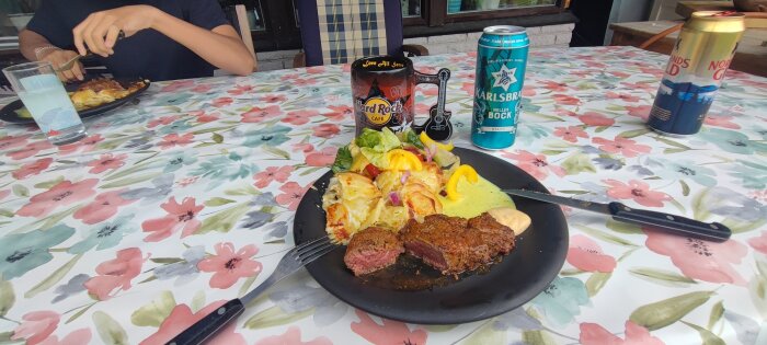 Matrester från grillmiddagen, inklusive kött, gratäng och sallad, serveras på en tallrik. Måltiden är uppdukad på en blommig duk tillsammans med olika drycker.