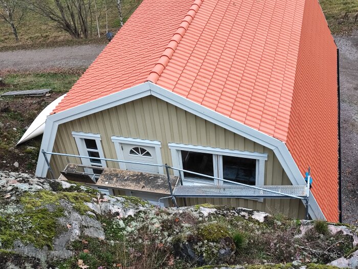 Garage med loft, gul träfasad och orange tegelrött tak, nära en bergvägg. Byggnadsställning vid ingången, mossig berggrund i förgrunden.