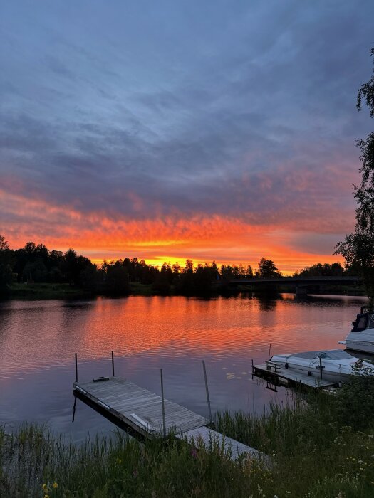 Solnedgång över en sjö med en brygga och båtar i förgrunden, omgiven av träd och med en himmel färgad i orange och lila toner.